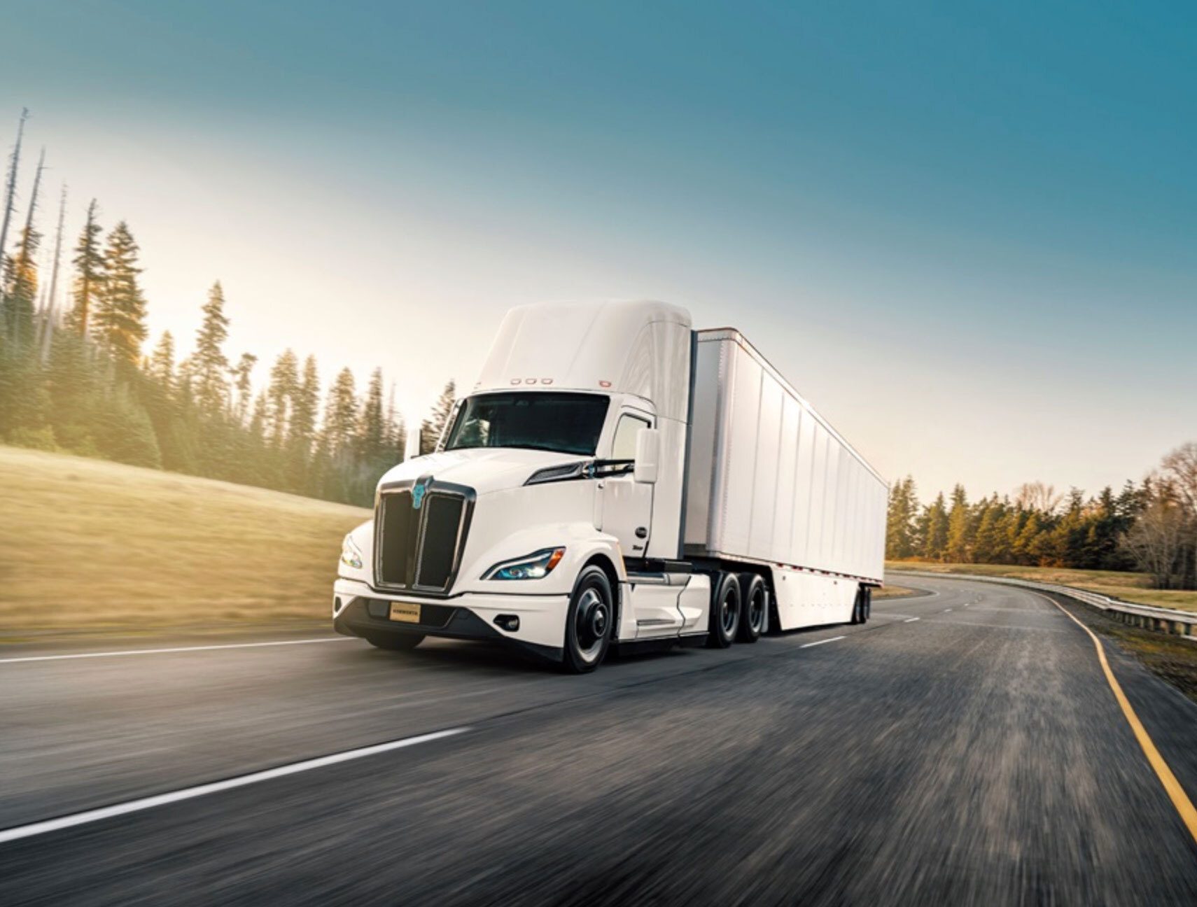 White semi-truck driving on a winding road among trees at sunset.