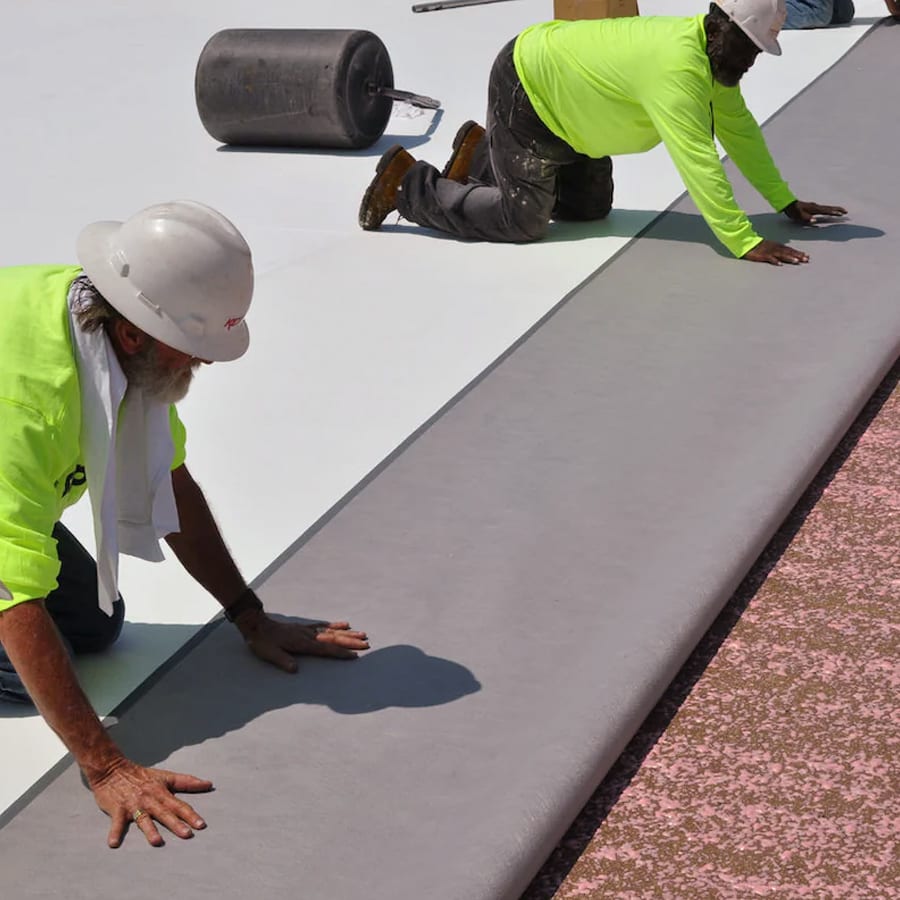 Two roofers in hardhats on their knees placing PVC membrane.