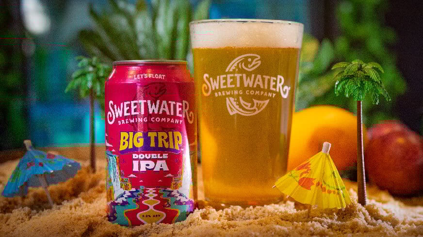 SweetWater Big Trip Double IPA can and glass on a miniature beach with palm trees and umbrellas.
