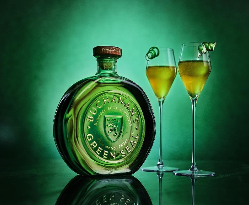 Buchanan's Green Seal whisky bottle and two green-hued cocktails with lime peels on a dark green background.