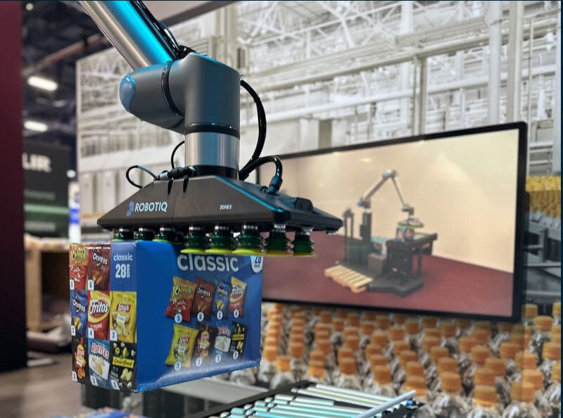 Robotic arm with a ROBOTIQ gripper holding a box of Classic snack bags in an industrial setting.