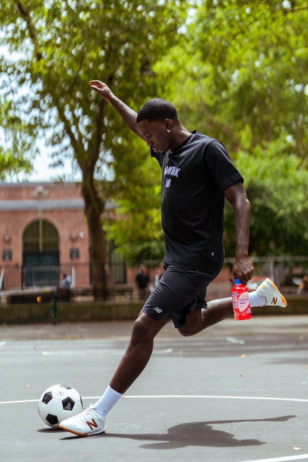 A man in athletic wear kicks a soccer ball on a court, holding a red drink.