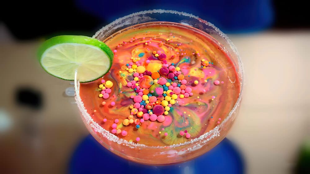 A vibrant rainbow drink with colorful sprinkles, a lime slice, and a sugar-frosted rim.