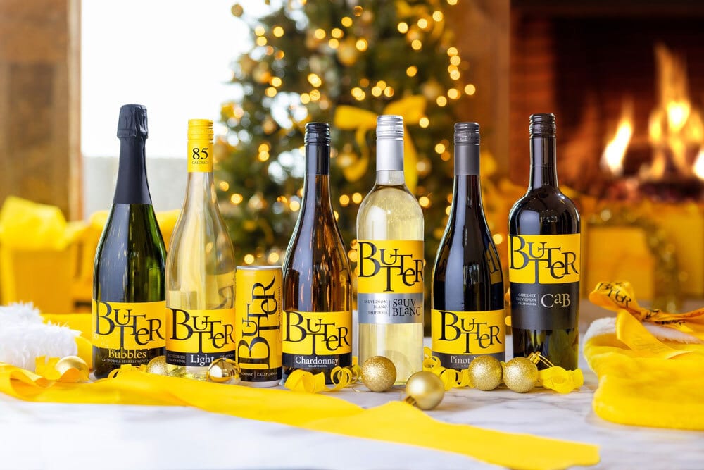 Assorted Butter brand wines (bottles, can) in a festive Christmas setting with tree & fireplace.