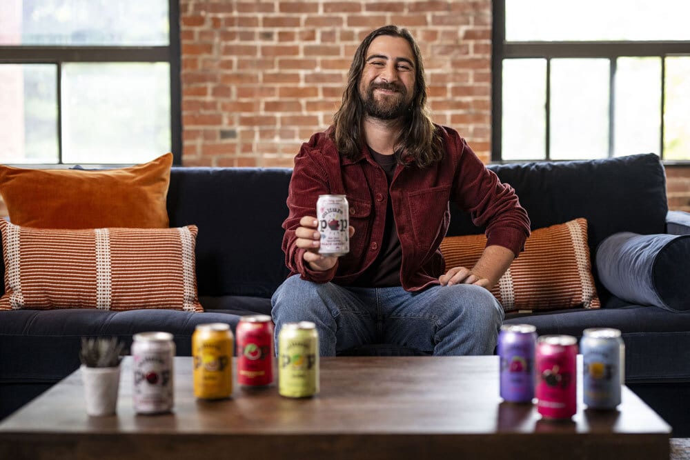 Man smiling on a couch, holding a drink can, with more cans on a coffee table.