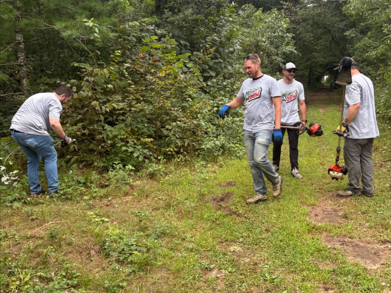 Four men in Mountain Dew shirts clearing an overgrown path in a natural, wooded setting.