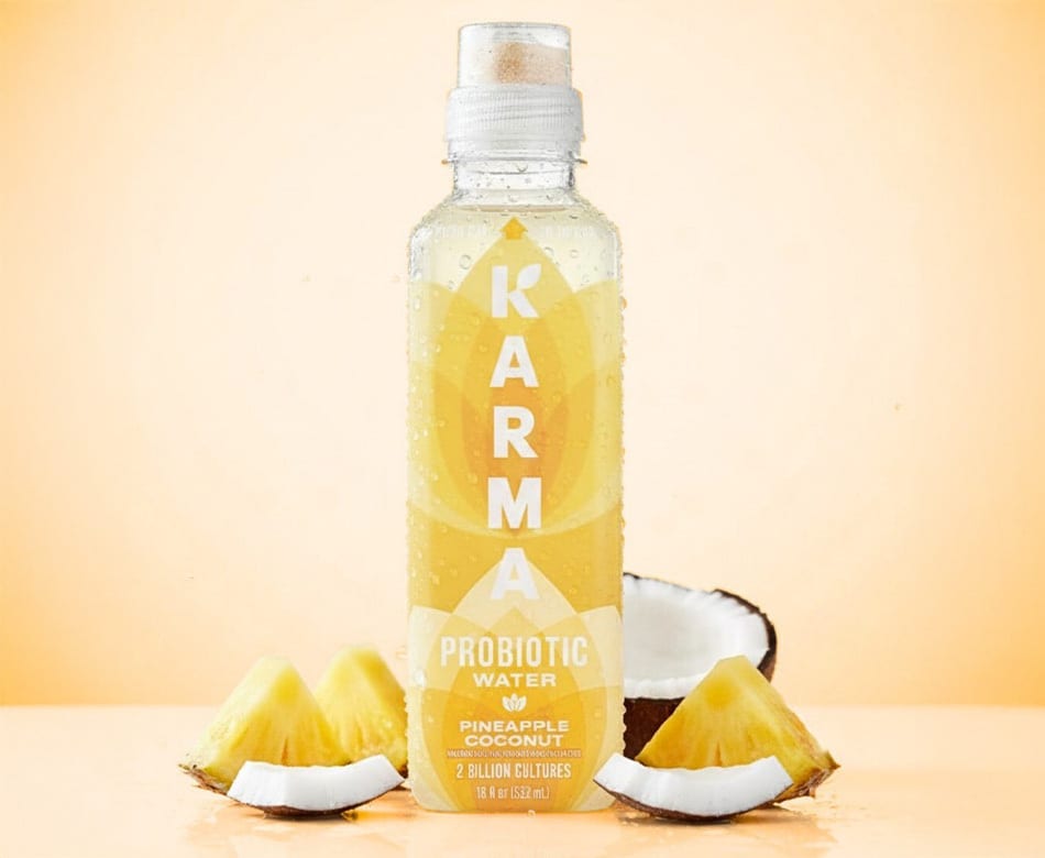 Karma Pineapple Coconut Probiotic Water bottle with fresh pineapple & coconut pieces on an orange background.