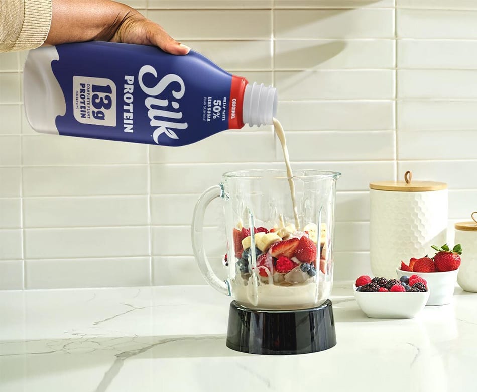 A hand pours Silk Protein milk into a blender filled with mixed fruit for a smoothie on a kitchen counter.