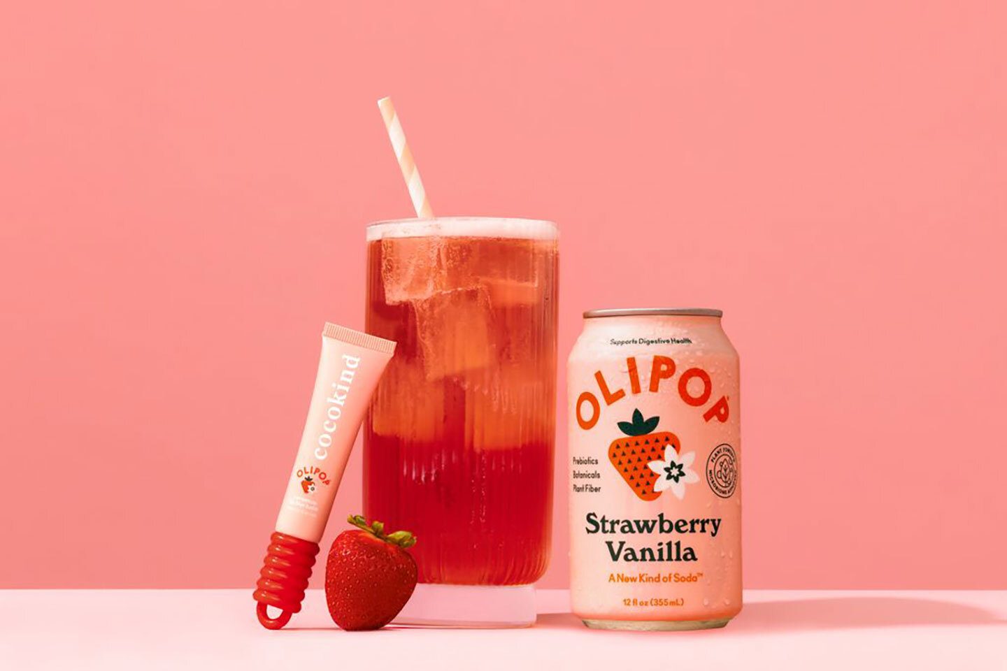 A pink OLIPOP Strawberry Vanilla soda can, glass with drink and straw, Cocokind tube, and a strawberry.