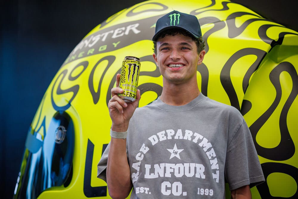 Smiling Lando Norris in a Monster Energy cap holds a Monster drink.