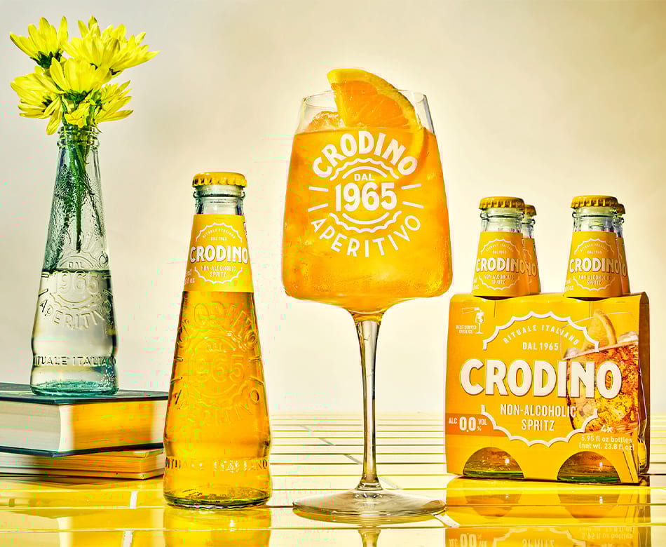 Crodo non-alcoholic aperitivo: glass with drink, bottles, 4-pack, flowers, books on yellow.