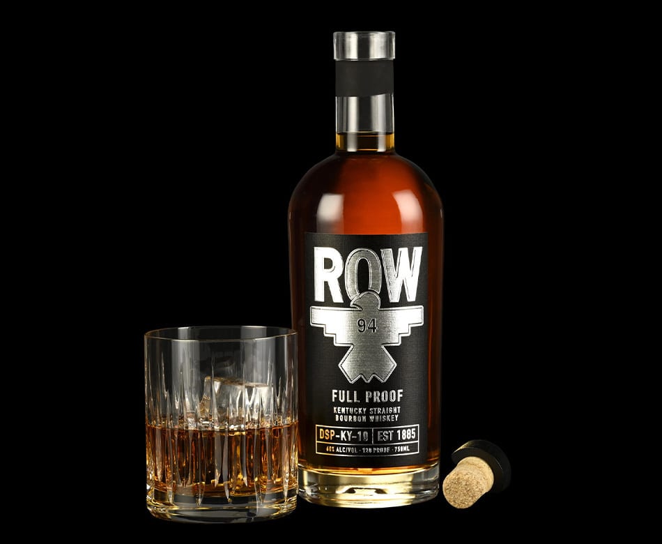 Bottle of ROW Full Proof Bourbon, a glass of whiskey with ice, and a cork on a black background.