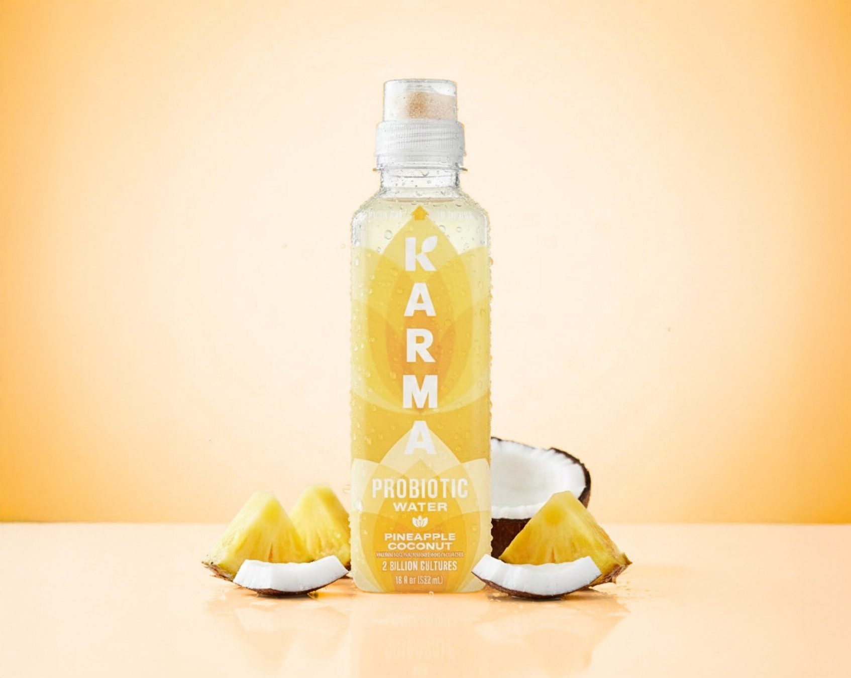 A bottle of Karma Pineapple Coconut Probiotic Water surrounded by fresh pineapple and coconut pieces on a warm orange background.