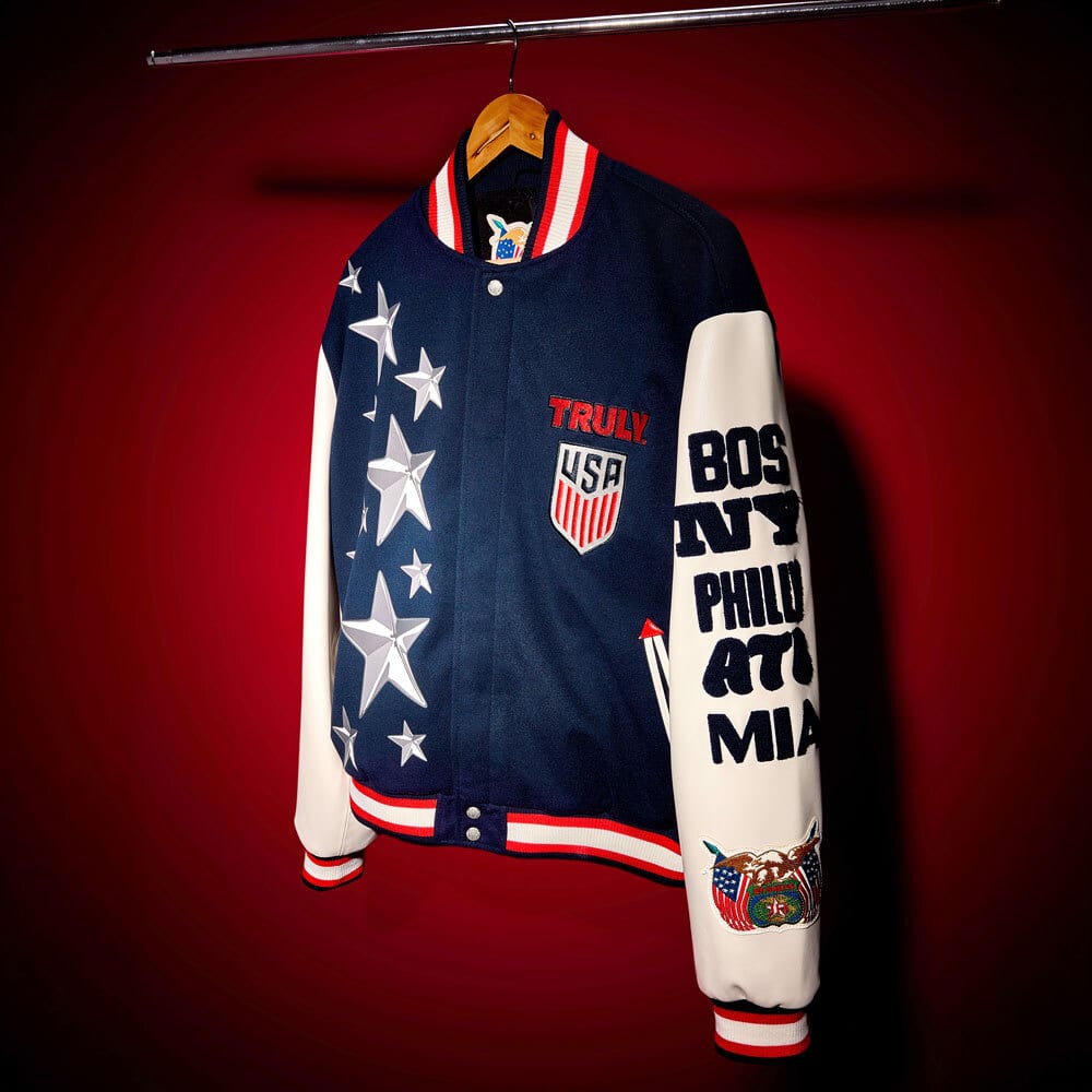 USA-themed navy and white varsity jacket with stars and city names on sleeve, hanging on a rack.