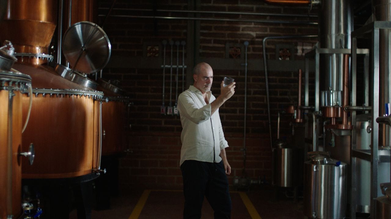 A man inspects a glass in a distillery with large copper stills.