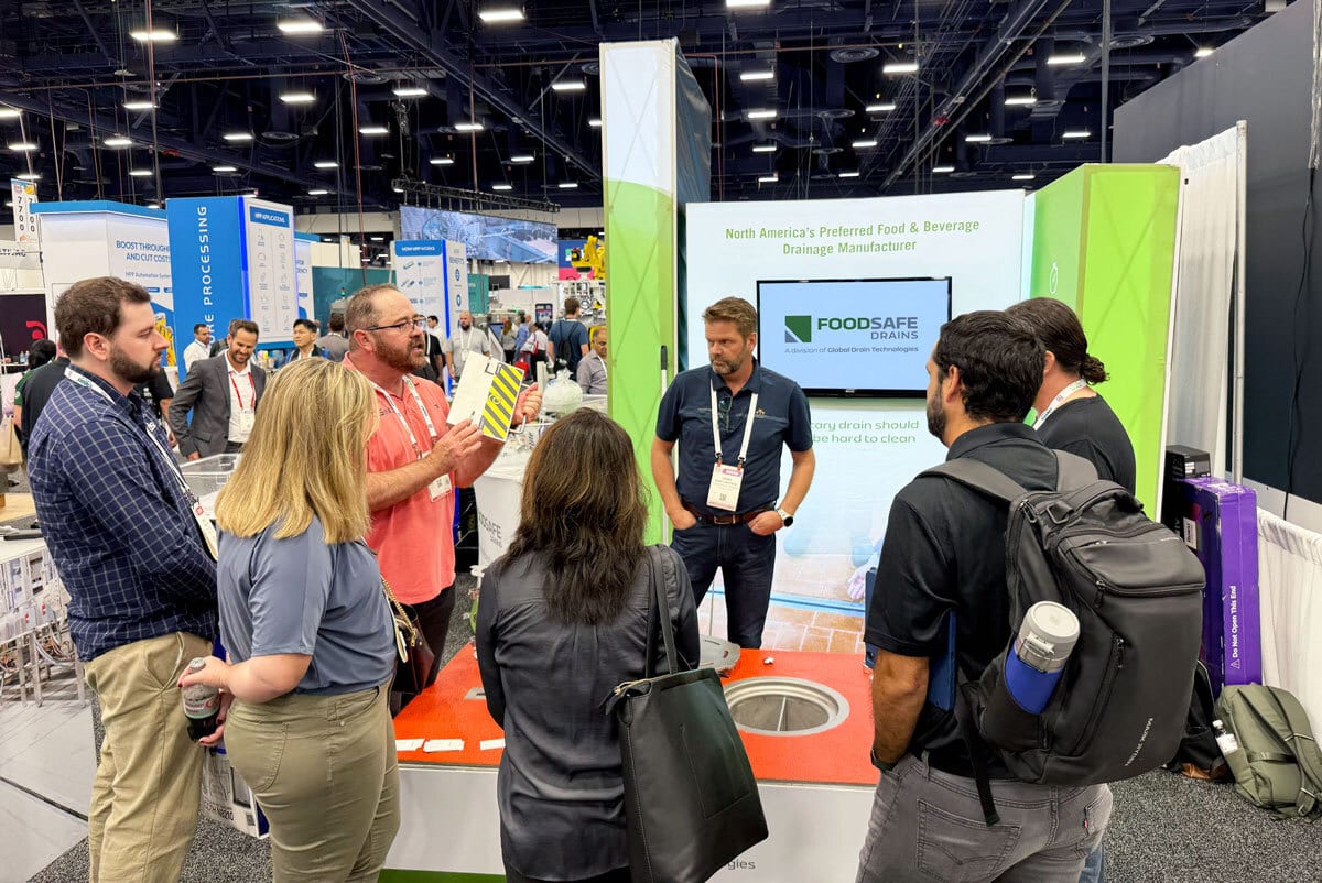 Attendees at a trade show booth engaging with a speaker about Foodsafe Drains, featuring a display model.