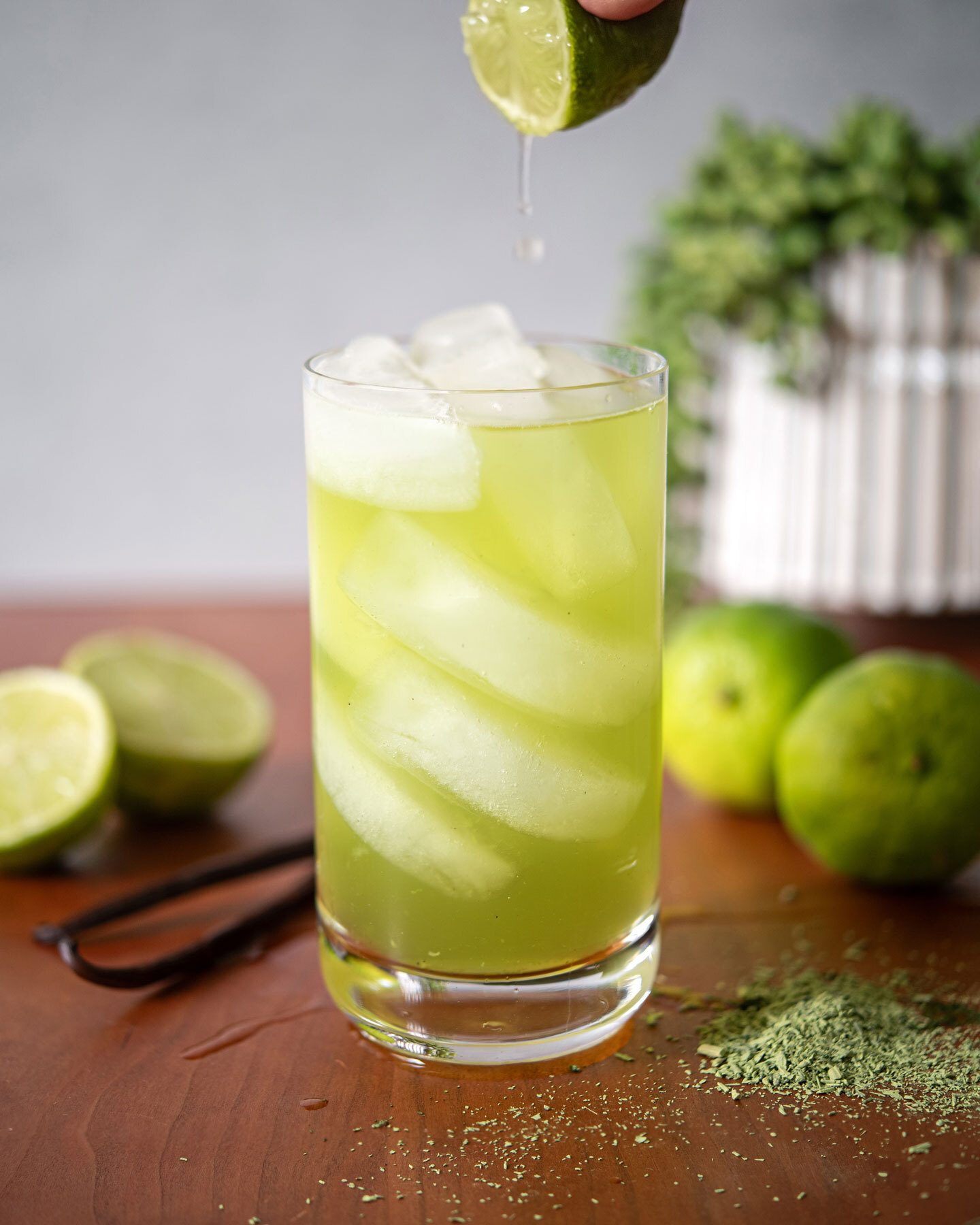 Lime squeezed into a glass of green iced drink. Limes, vanilla beans, and matcha on wood.
