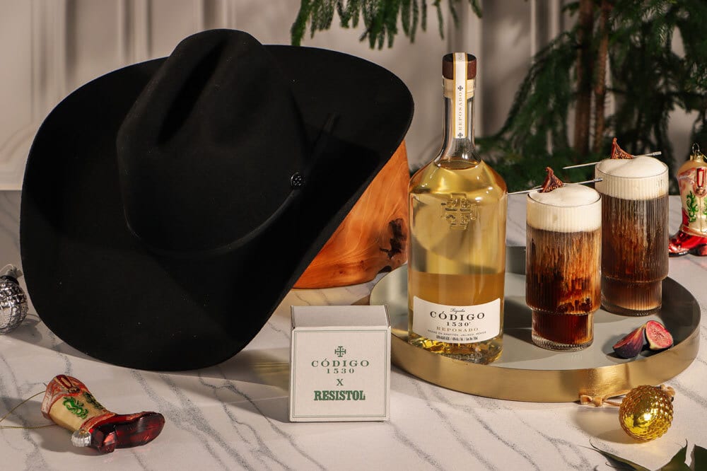 A black cowboy hat, Código 1530 tequila bottle, two cocktails, and festive holiday decorations on a tray.