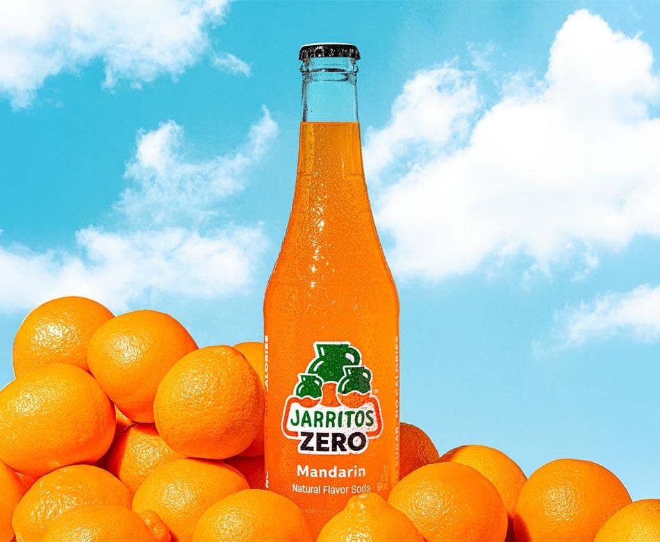 Jarritos Zero Mandarin soda bottle surrounded by mandarins, against a blue sky with clouds.