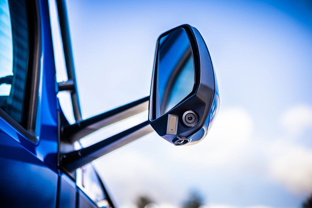 Blue vehicle side mirror with integrated camera and sensor, against a bright sky.