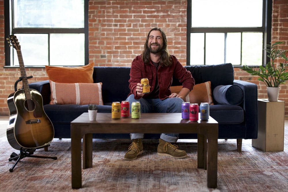Smiling man on couch holds a can; colorful cans on table, guitar, brick wall background.