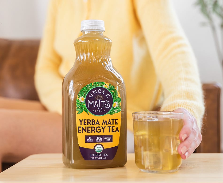 A large bottle of Uncle Matt's Organic Yerba Mate Energy Tea next to a glass of the tea, held by a person.