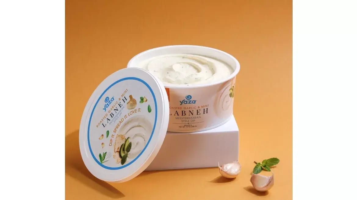 Open tub of Yaza Roasted Garlic & Mint Labneh dip with lid, garlic cloves, and mint leaves.