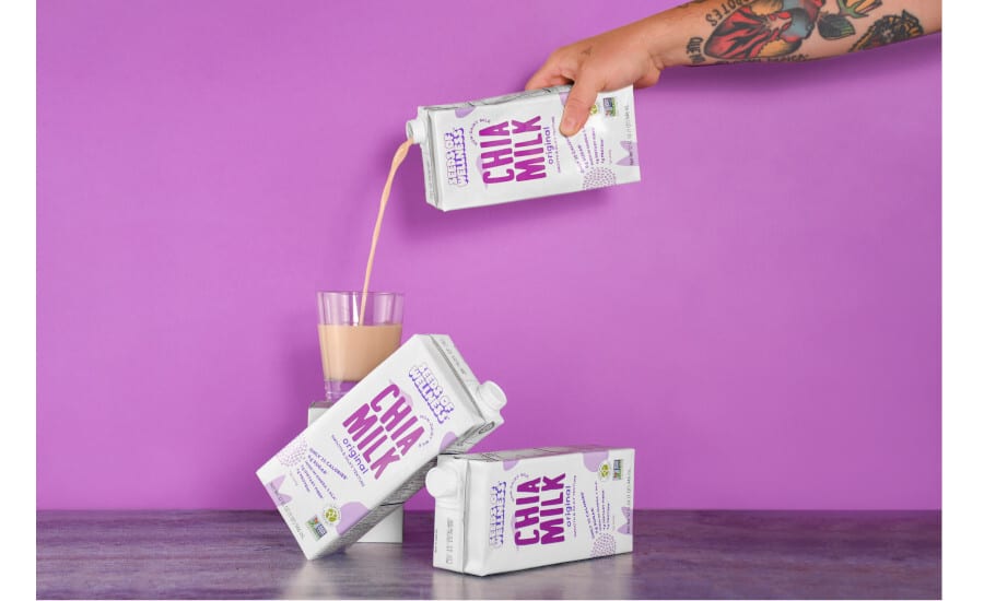 Hand pours beige Chia Milk from carton into glass. Two cartons on surface below. Purple background.