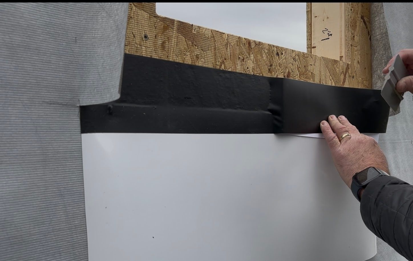 Hands applying black flashing tape below a window opening on a building exterior.
