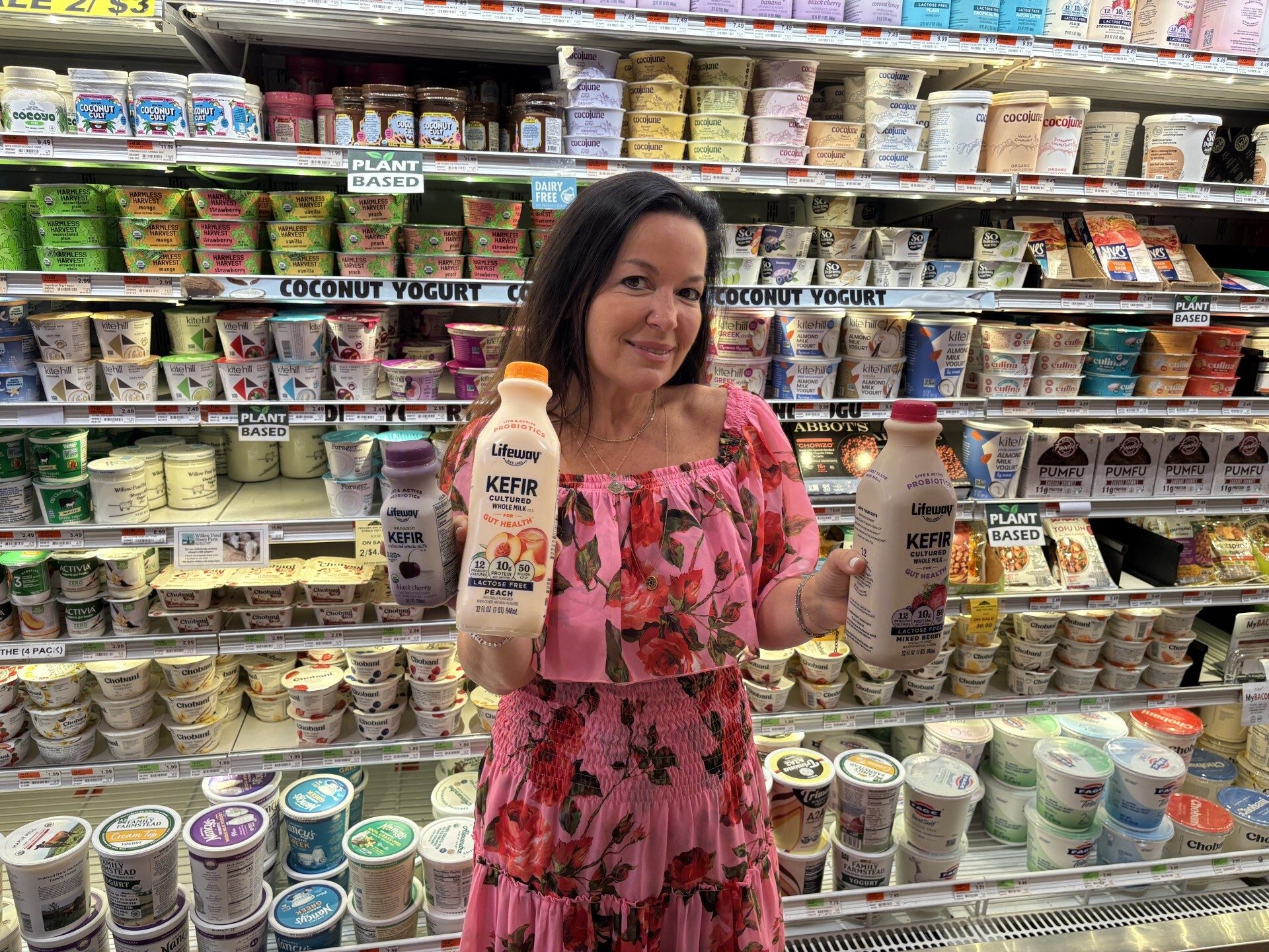 Woman holds two Lifeway kefir bottles in a grocery store aisle filled with yogurts and plant-based dairy.