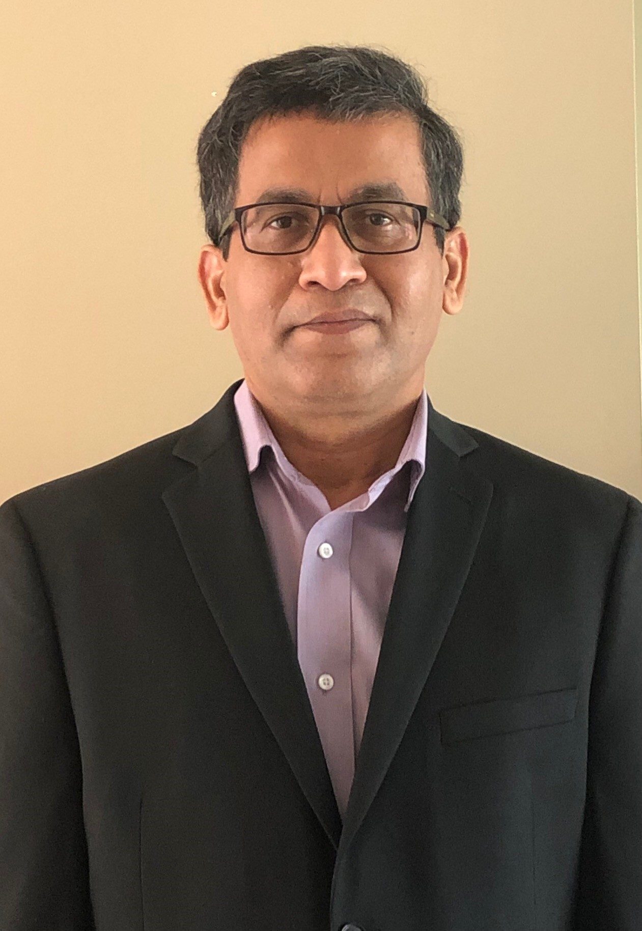 Headshot of a man wearing a black suit, lavender shirt, and black-framed glasses.