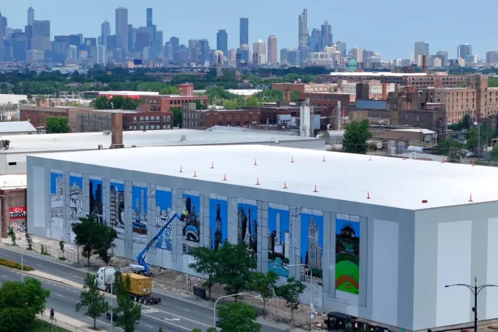 Large building with Chicago mural being painted, distant city skyline behind it.
