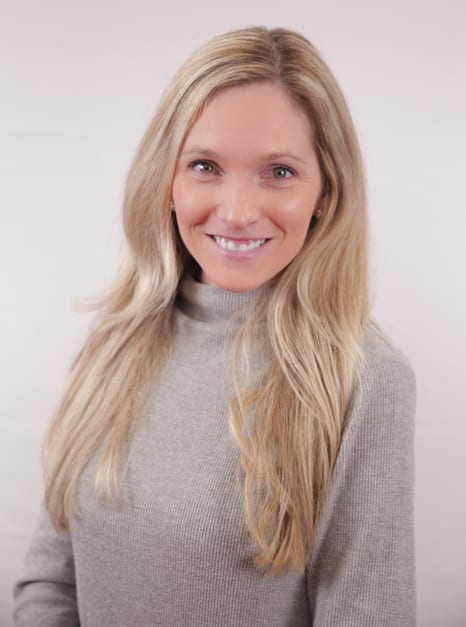 A headshot of a smiling woman with long blonde hair wearing a grey turtleneck sweater against a light background.