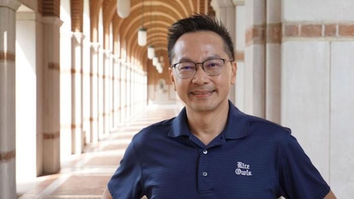 Smiling man in glasses wearing a "Rice Owls" polo shirt in an arched hallway.