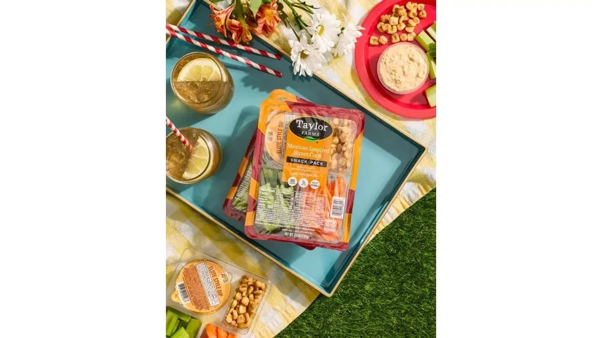 Picnic spread featuring Taylor Farms Mexican Street Corn snack packs, drinks, dip, veggies, and flowers.