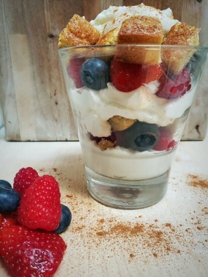 A clear glass filled with layered berries, cream, and cake, topped with cinnamon.