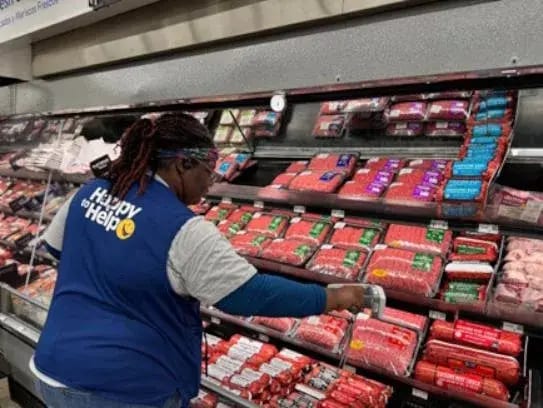 "Happy to Help" employee in a blue vest scanning meat packages in a grocery store display.