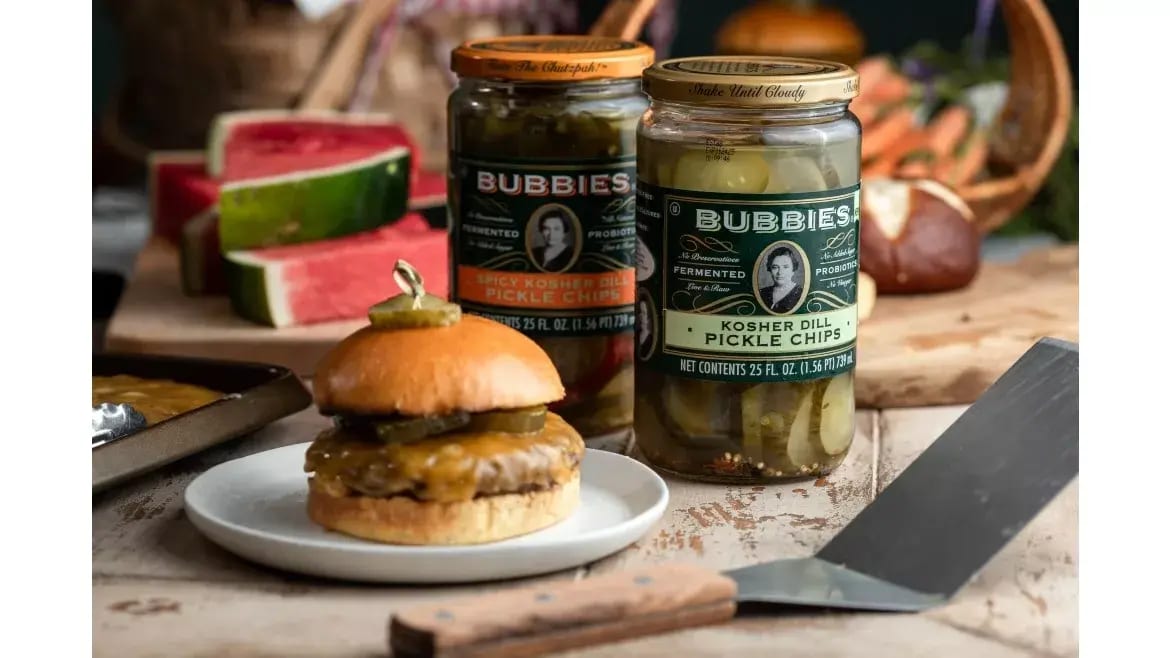 Two jars of Bubbies pickles, a cheeseburger, and watermelon slices on a wooden surface.