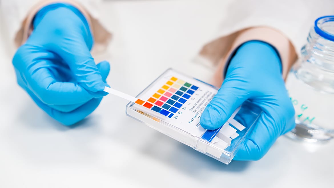 Gloved hands holding a pH test strip and comparing it to a pH color chart.