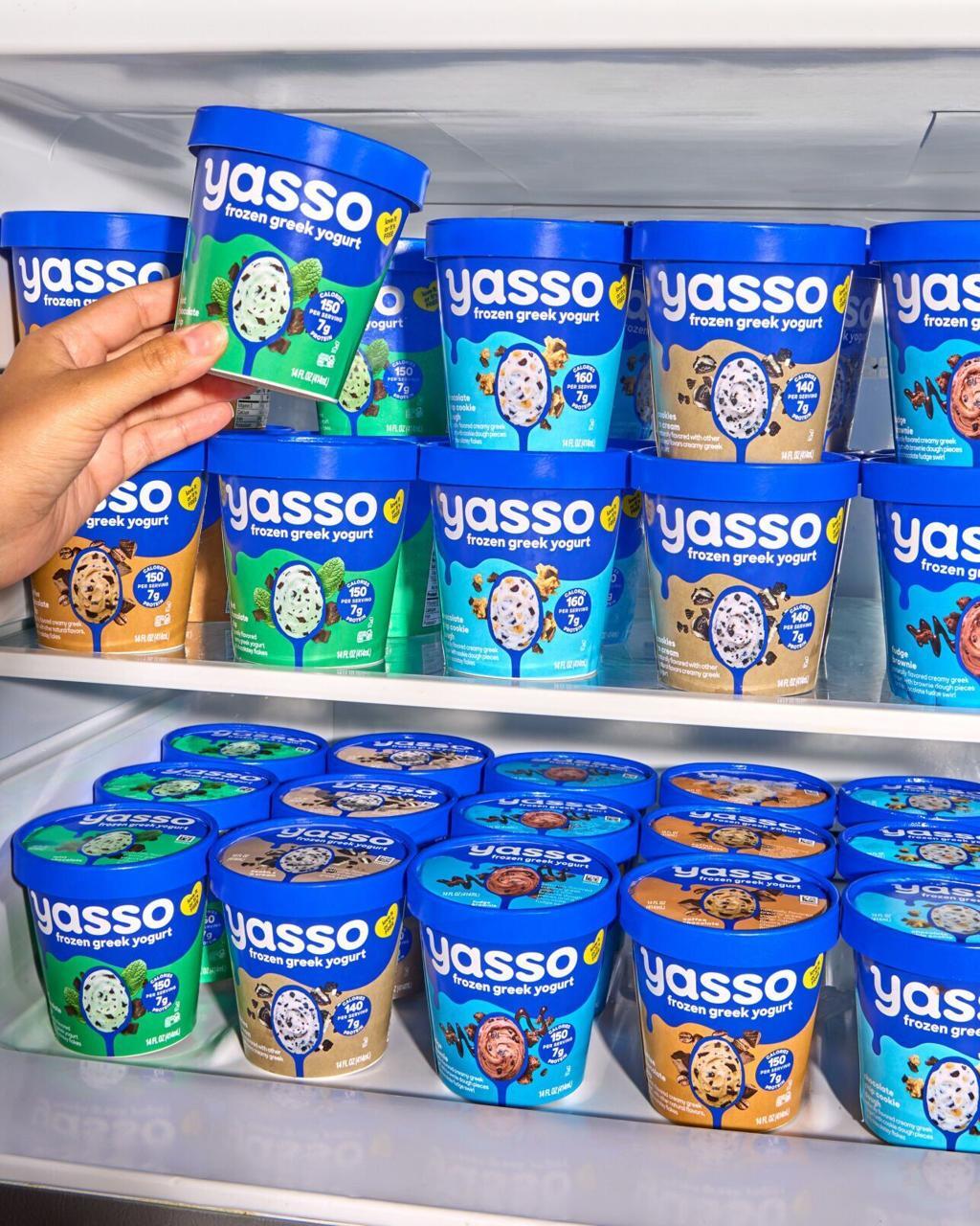 A hand selects a Yasso frozen Greek yogurt tub from a freezer full of various flavors.