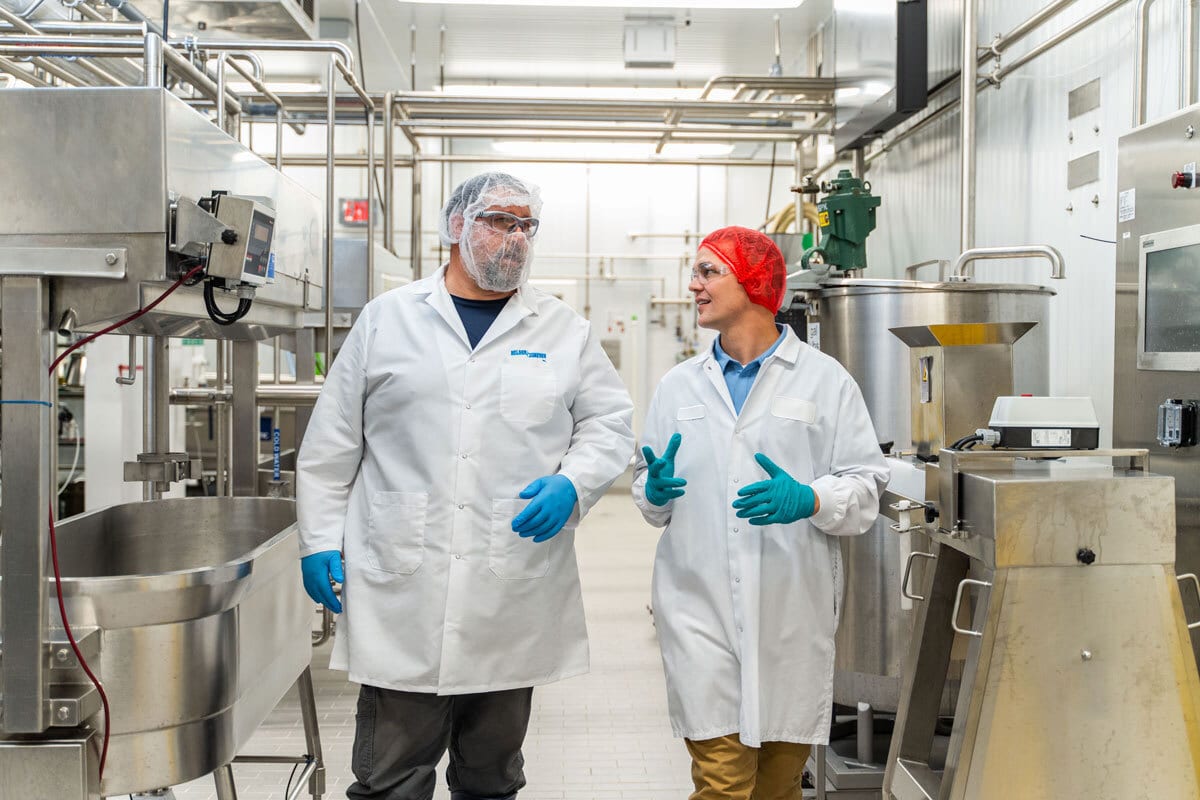 Two people in lab coats, hairnets, and gloves discussing in a stainless steel processing facility.