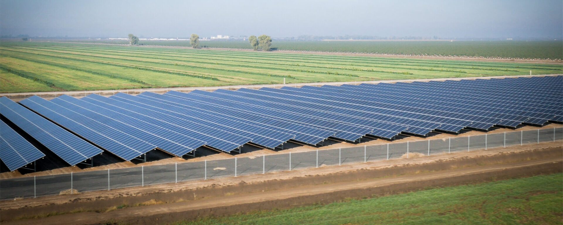 Aerial view of a large solar panel farm next to agricultural fields under a hazy sky.