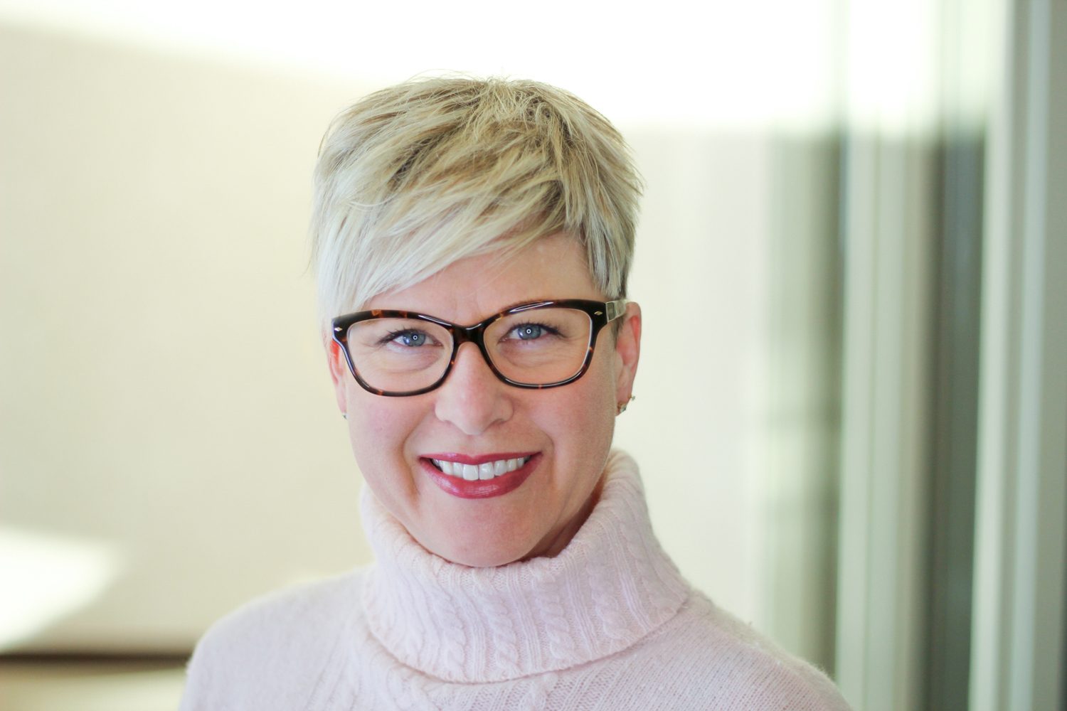 A woman with short blonde hair and glasses smiles directly at the camera, wearing a light pink turtleneck.