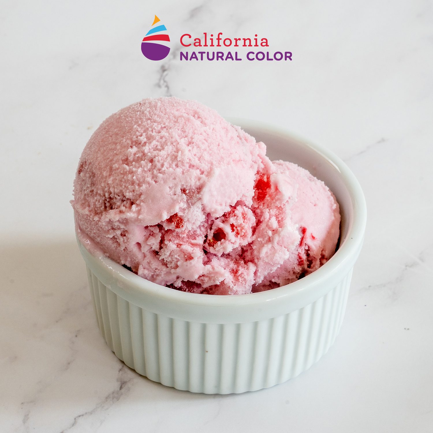 Two scoops of pink strawberry ice cream in a white ceramic ramekin on a white marble surface.