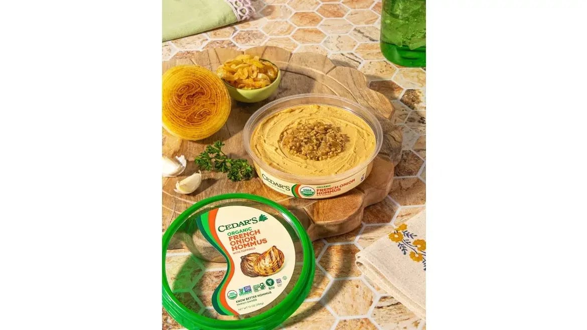 Two Cedar's Organic French Onion Hommus tubs, one open with toppings, the other closed, on a board.