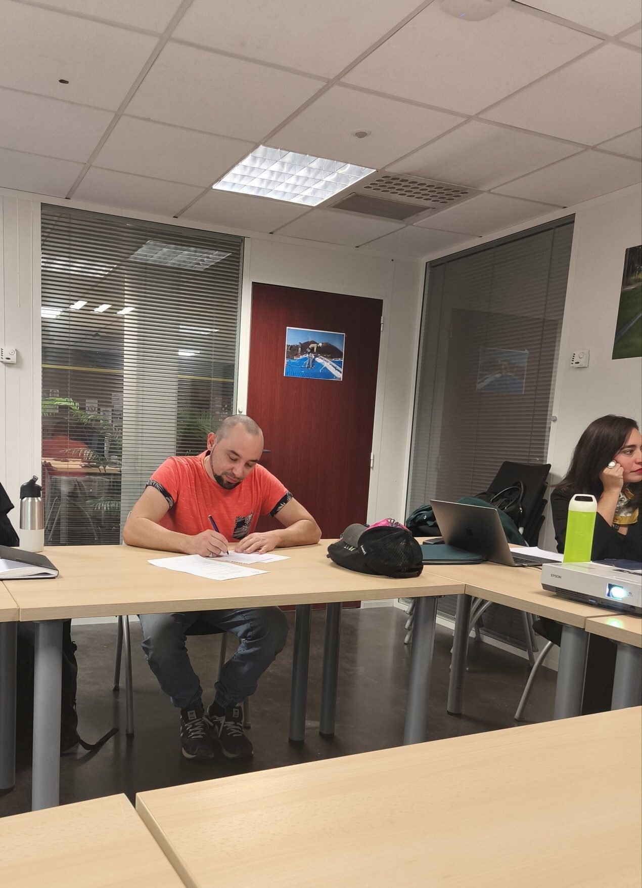 A man writes at a table in a brightly lit modern meeting room, with another person visible.