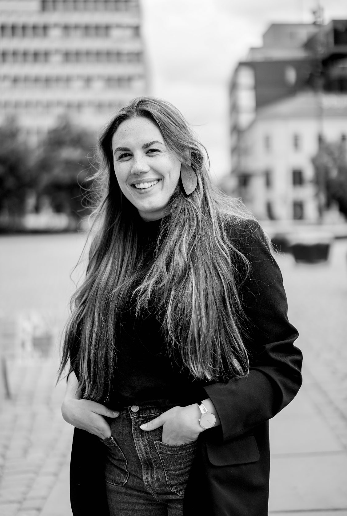 B&W smiling woman with long hair, hands in pockets, city background.