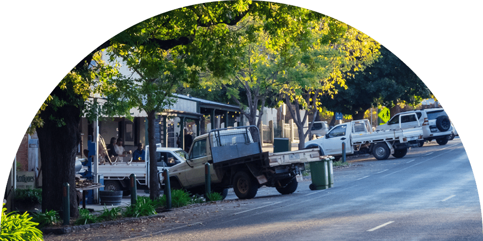 A sunlit street lined with parked pickup trucks, vibrant green trees, and people relaxing outside a cafe.