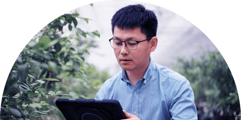 An Asian man in glasses and a blue shirt uses a tablet among green plants, likely for agricultural work.