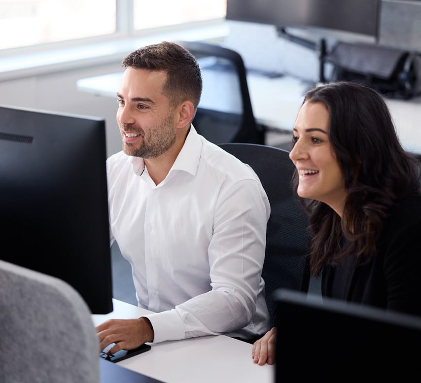 Two smiling colleagues, a man and a woman, collaborate at a computer in a modern office.